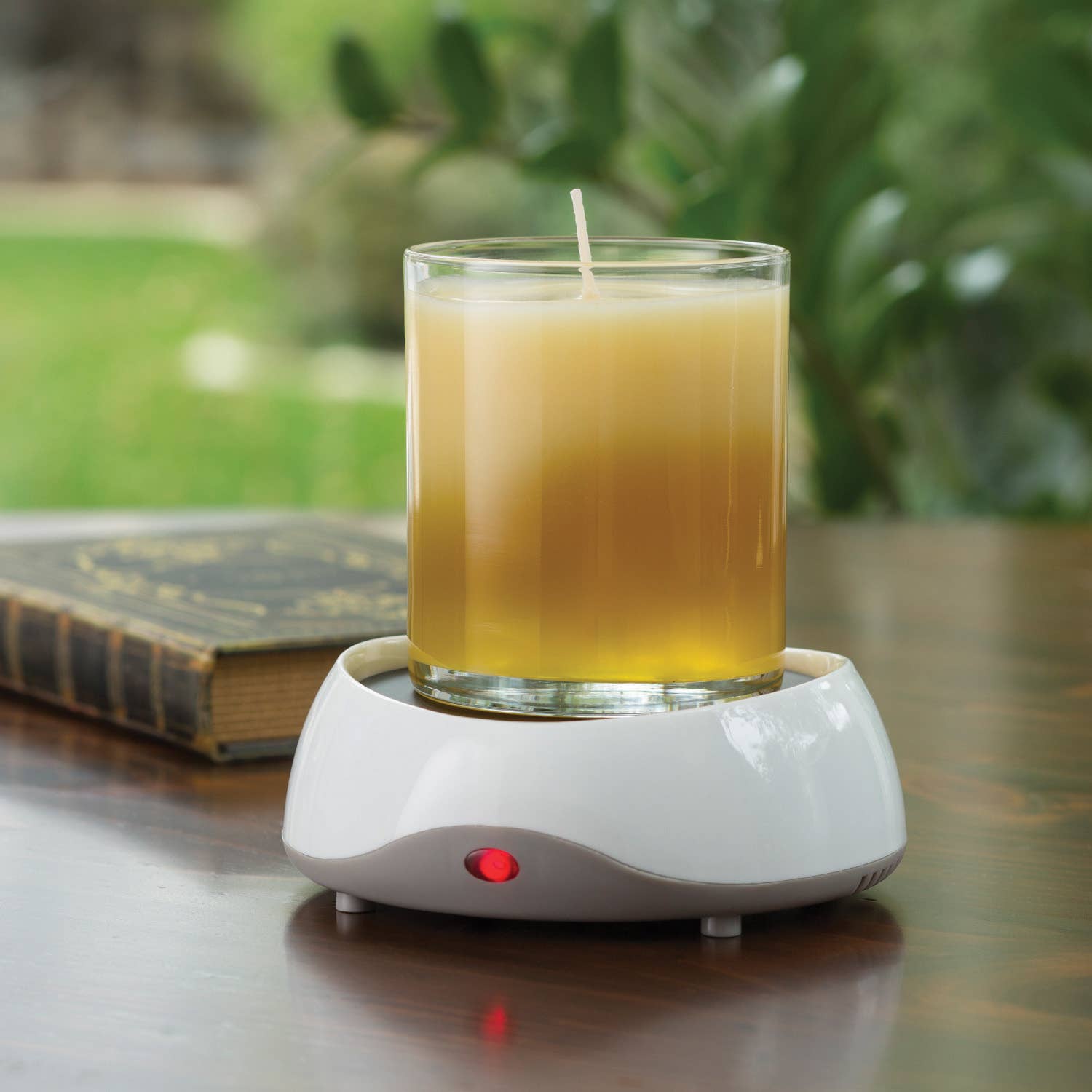 Candle in a glass holder on a white candle warmer with a blurred green outdoor background