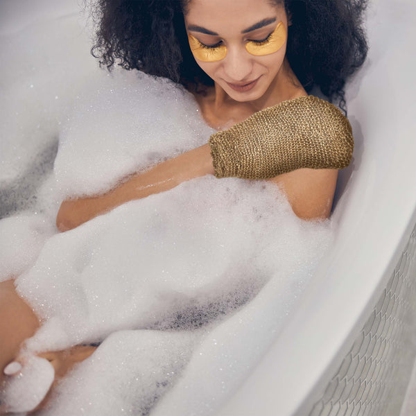 Woman in a bathtub with bubbles, wearing gold gloves and face masks.